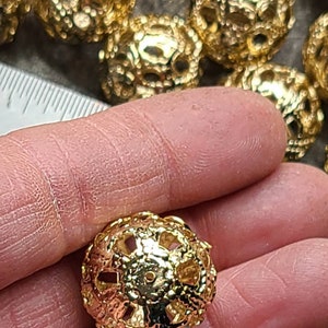May include: A close-up of a gold-colored metal bead with a decorative, openwork design. The bead is round and has a slightly textured surface.