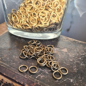 May include: A glass jar filled with many small, gold-coloured metal rings. A small pile of the same rings sits outside the jar on a dark, textured surface. The rings are open jump rings, suitable for jewellery making or crafts.