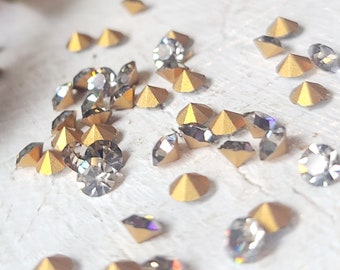 May include: Assortment of gold and clear rhinestones, scattered on a white surface. The rhinestones have a faceted, pointed design.