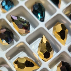 May include: A white plastic tray with compartments filled with various iridescent glass teardrop-shaped beads. The beads are in shades of gold, blue, and purple.