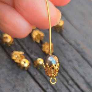 May include: Close-up of a gold-colored jewelry component with a dark gray pearl bead. The bead is set in a decorative gold-colored cap. A gold wire extends from the bead, and other similar components are scattered in the background.
