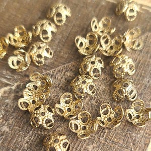 May include: A collection of gold-toned metal bead caps with a decorative, openwork design. The caps are scattered on a wooden surface.