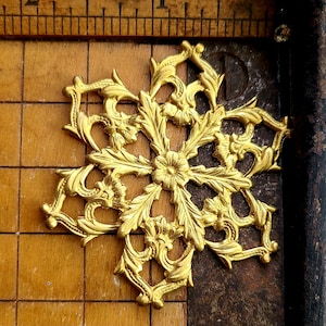 May include: A gold-colored, ornate metal embellishment in a floral snowflake design. The decorative piece features detailed leaf and scrollwork patterns. The object is set against a wooden background with a ruler.