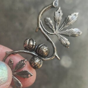 May include: A silver-toned metal charm featuring a stylised vine with leaves and berries. The charm is detailed with intricate carvings and has a rustic, antique look.
