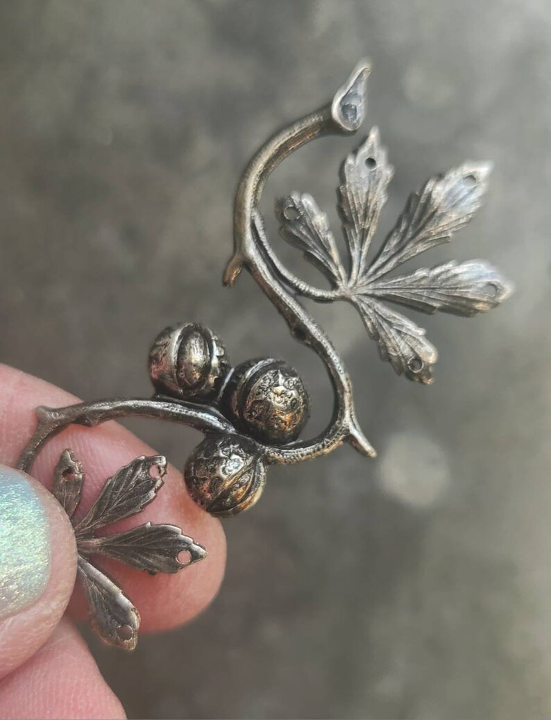 May include: A silver-toned metal charm featuring a stylised vine with leaves and berries. The charm is detailed with intricate carvings and has a rustic, antique look.