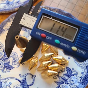 May include: A collection of gold-colored, cone-shaped metal studs with a textured surface, displayed on a blue and white floral patterned plate. A digital caliper measures one stud at 1.14 mm. The studs are small and pointed.