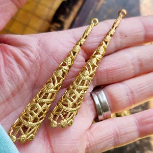 May include: Two gold-tone metal filigree pendant charms in the shape of a long, narrow cone. The charms have a delicate, intricate design and are perfect for adding a touch of elegance to jewelry projects.