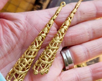 May include: Two gold-tone metal filigree pendant charms in the shape of a long, narrow cone. The charms have a delicate, intricate design and are perfect for adding a touch of elegance to jewelry projects.