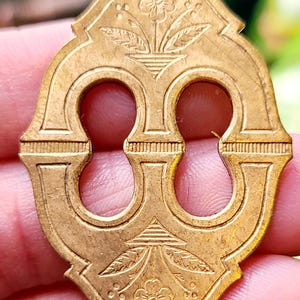 May include: A gold-colored metal keyhole cover with intricate floral designs. The cover has two keyhole openings and a decorative pattern around the edges.
