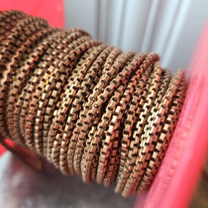 May include: Close-up of a spool of gold-colored chain. The chain is tightly wound, revealing a detailed pattern of rectangular links. The spool is set against a red and white background, highlighting the chain's metallic sheen.