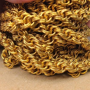 May include: Close-up of a gold-colored chain with a woven design. The chain is made of multiple interlocking links, creating a textured and ornate appearance. The chain is likely jewelry, such as a necklace or bracelet.