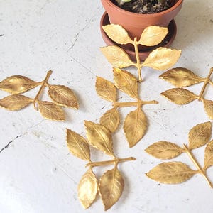 May include: Several gold-colored metal leaf sprigs are arranged on a white surface. Each sprig has multiple detailed leaves. A small potted plant is in the background. The leaves have a metallic finish.
