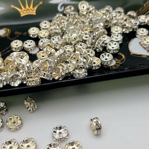 May include: A large pile of silver-toned metal bead caps with clear rhinestones. The bead caps are scattered on a black tray with a gold crown design. Some bead caps are also scattered on a white surface.