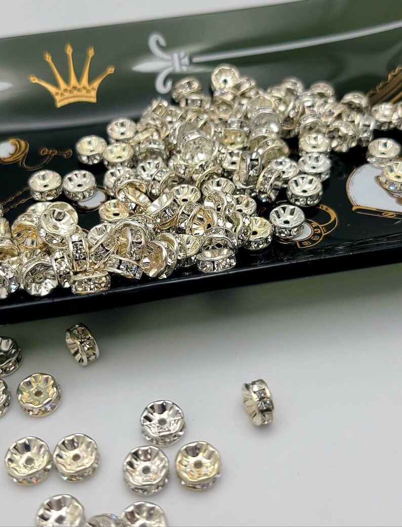 May include: A large pile of silver-toned metal bead caps with clear rhinestones. The bead caps are scattered on a black tray with a gold crown design. Some bead caps are also scattered on a white surface.