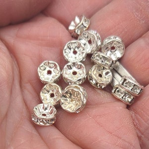 May include: A close-up of a hand holding a group of silver-toned metal bead caps with clear rhinestones. The bead caps have a decorative, scalloped edge.