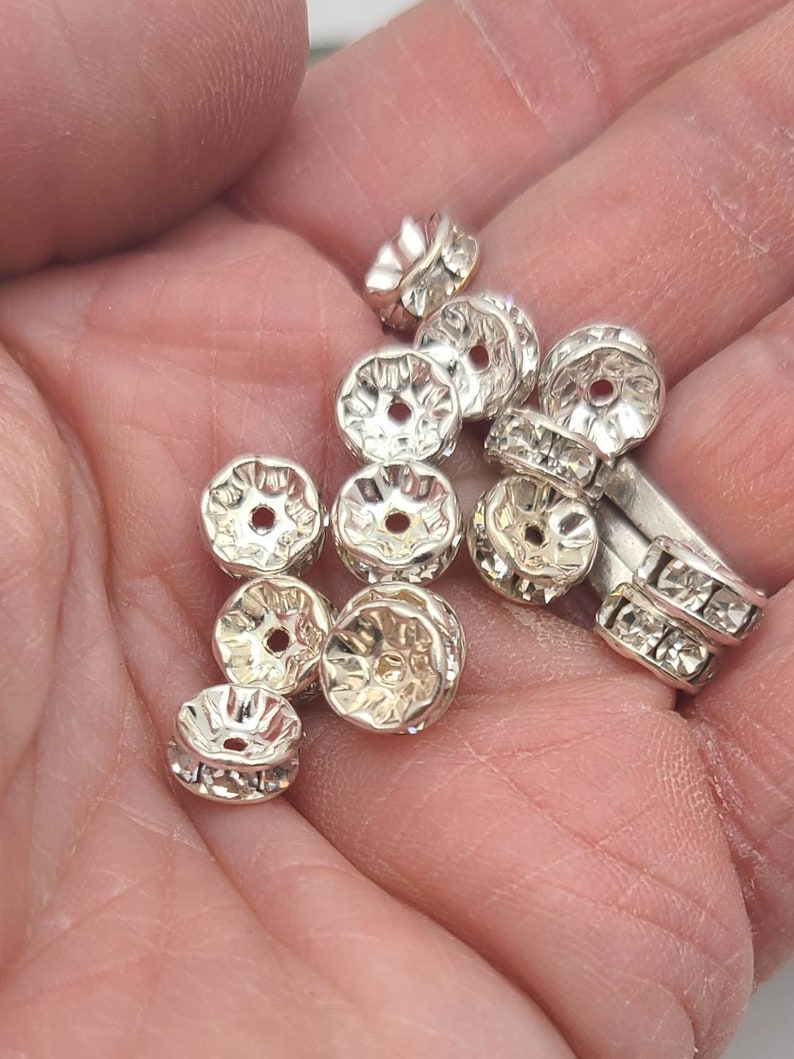 May include: A close-up of a hand holding a group of silver-toned metal bead caps with clear rhinestones. The bead caps have a decorative, scalloped edge.
