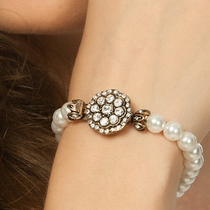 May include: A pearl bracelet with a decorative, round, antique gold-toned clasp. The clasp is adorned with clear, sparkling crystals. The bracelet features multiple white, round pearls, creating an elegant and classic look.