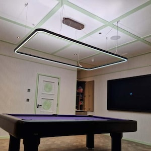 Può includere: Un tavolo da biliardo rettangolare nero con un piano in feltro viola è al centro di una stanza. Una grande lampada rettangolare nera è appesa sopra. Le pareti sono chiare con strisce verticali e un grande schermo nero è montato sulla parete.