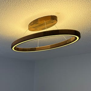 May include: Contemporary wooden oval chandelier with a warm, glowing light. The fixture is suspended from the ceiling and features a smaller, matching wooden disc above. The design is minimalist and modern, suitable for home decor.