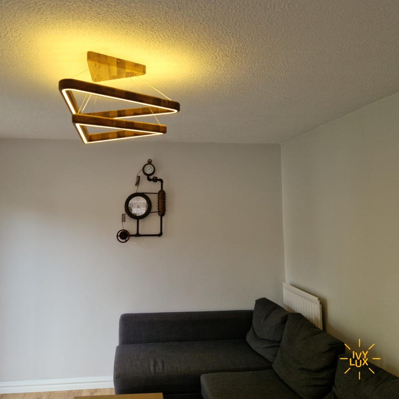 May include: A modern wooden triangle chandelier with three tiers hangs from the ceiling in a living room. The chandelier is lit with warm white LED lights. A gray couch is in the foreground, and a steampunk-style clock is on the wall.