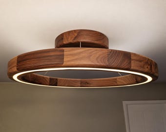 May include: A circular flush mount ceiling light fixture constructed from segments of dark brown walnut wood. The fixture features a central circular base and a larger outer ring with an integrated LED light strip emitting a warm glow.