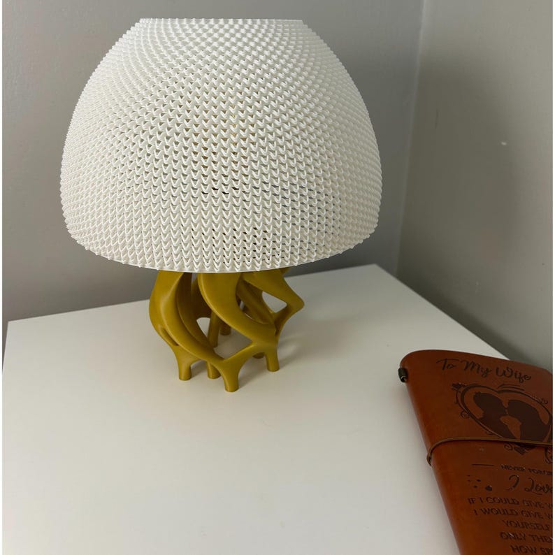 May include: A white lampshade with a textured pattern sits atop a gold, abstract, 3D printed base. The lamp is on a white surface next to a brown leather journal with the words "To My Wife" embossed on the cover.