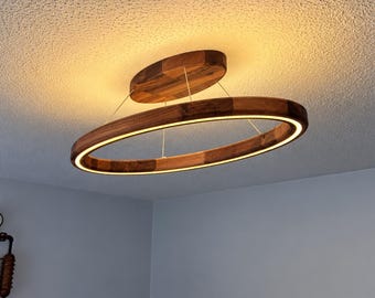 May include: An oval-shaped wooden ceiling light fixture with a natural grain finish. The fixture features a central wooden mount attached to the ceiling, with a larger oval wooden ring suspended below by thin wires, emitting a warm internal glow.