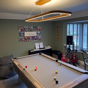 Può includere: Un apparecchio di illuminazione rettangolare in legno con una striscia di luce LED bianca è appeso al soffitto sopra un tavolo da biliardo. Il tavolo da biliardo ha una superficie in feltro grigio ed è circondato da una cornice in legno. Ci sono diverse palle da biliardo sul tavolo, tra cui palle rosse, gialle e bianche. La stanza ha pareti verdi e un pavimento in legno. C'è un cartello sulla parete che dice "PLAY".