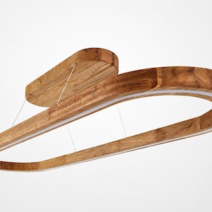 Puede incluir: Una lámpara colgante con un marco de madera curvado de forma ovalada y veta natural visible. Una tira de luz LED blanca integrada recorre el borde interior de la madera, suspendida por cables finos desde un soporte de techo de madera a juego.