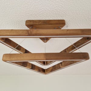 May include: A modern wooden ceiling light fixture with a geometric design. The light is made of two square frames, one inside the other, creating a triangular shape. The light is suspended from the ceiling by wires.