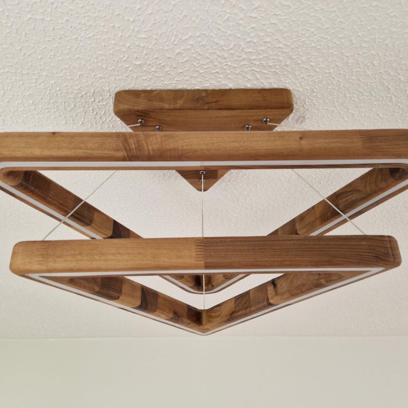 May include: A modern wooden ceiling light fixture with a geometric design. The light is made of two square frames, one inside the other, creating a triangular shape. The light is suspended from the ceiling by wires.