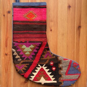 May include: A patterned Christmas stocking with a geometric design. The top section is pink with a diamond pattern, followed by brown and red stripes. The lower section features red, purple, and brown geometric shapes. A blue loop is at the top.