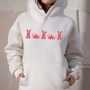 Op de afbeelding: Witte hoodie met een roze geborduurd ontwerp van konijntjes met strikjes.