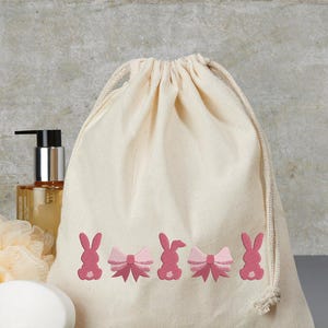 Op de afbeelding: Een witte katoenen trekkoordtas met roze geborduurde konijntjes en strikken. De tas ligt op een grijs oppervlak met een fles lotion, een stuk zeep en een washandje.