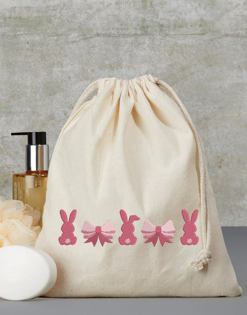 Op de afbeelding: Een witte katoenen trekkoordtas met roze geborduurde konijntjes en strikken. De tas ligt op een grijs oppervlak met een fles lotion, een stuk zeep en een washandje.