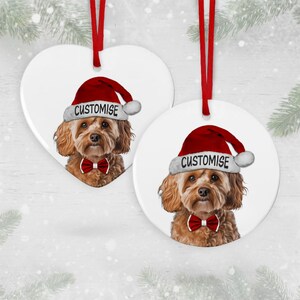 May include: Two white ceramic Christmas baubles featuring a brown and white dog wearing a red Santa hat and a red bow tie. The text "CUSTOMISE" is on the hat of each bauble.