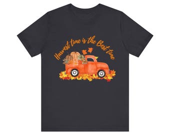 Puede incluir: Una camiseta gris oscuro con el texto "Harvest time is the best time" en letras naranjas. Debajo del texto se ve una camioneta roja con la caja llena de calabazas y hojas de otoño.