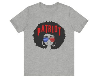 Peut inclure: T-shirt gris avec une illustration en noir et blanc d'une femme avec une afro portant des lunettes de soleil avec le motif du drapeau américain. Le mot "PATRIOT" est écrit en lettres rouges au-dessus de la tête de la femme.
