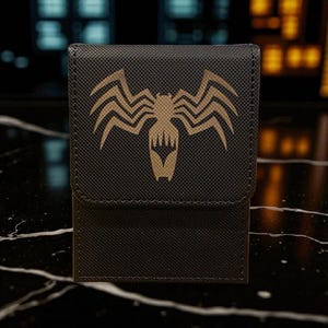 A black wallet or card case with a gold-colored spider logo on it, placed on a marble surface.