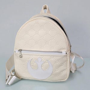 May include: Cream-colored backpack with a silver zipper and white straps. The front features a white Rebel Alliance symbol. The bag has a quilted pattern with repeating circular designs.