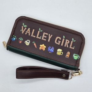 May include: A dark brown rectangular zip-around wallet with a dark green trim and a matching detachable wrist strap. The front features the text VALLEY GIRL in a light brown font, accented by small green leaves and various pixelated icons.