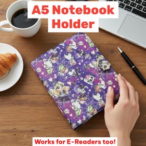 May include: A5 notebook holder with a purple and white pattern featuring cartoon characters, pumpkins, and spiderwebs. The holder has a button closure. Text on the image reads "A5 Notebook Holder" and "Works for E-Readers too!"