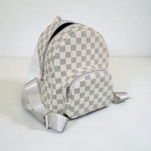 May include: A small, white and gray checkered backpack with a zippered main compartment and a smaller front pocket. The backpack has silver-colored zippers and adjustable white straps. The bag is made of a durable material and is designed for everyday use.