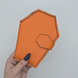 May include: An orange leather wallet with a unique geometric shape and black stitching. The wallet has a small, hexagonal flap with a matching orange strap for closure. The wallet is held in a hand against a white background.