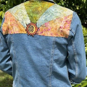 May include: A light blue denim jacket with a colorful patchwork design on the upper back. The patchwork features floral and abstract patterns in shades of orange, yellow, green, and blue. A sun-shaped patch with the text "You Are My Sunshine" is attached.