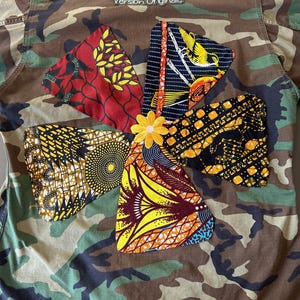 May include: Camouflage jacket with a floral design made of colorful African fabric patches. The patches are red, yellow, orange, and blue. The text "Version Originale" is above the floral design.