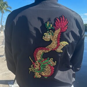 May include: Black bomber jacket with a large, embroidered dragon design on the back. The dragon features red, gold, and green detailing. The jacket is worn by a person, with the back of the jacket facing the camera.