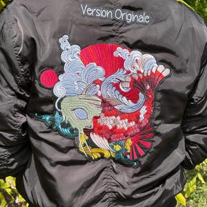 May include: Black bomber jacket with a colorful embroidered koi fish design on the back. The koi fish features red, white, and blue accents. The text "Version Originale" is embroidered on the upper back.