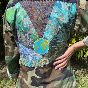 May include: Camouflage jacket with a patchwork back featuring floral and abstract patterns in shades of green, blue, and brown. A circular patch of the Earth is centered on the back. The text "Version Originale" is above the patchwork.