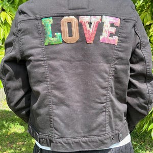 May include: Black denim jacket with a black faux fur collar. The word "LOVE" is spelled out across the back in colorful sequined letters. The jacket has button closures and pockets.